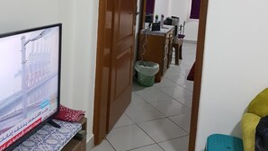 Ein Zimmer mit Fernseher auf einer Holzkommode, einem grünen Kunststoffhocker und einer Holztür, die in einen anderen Raum führt.