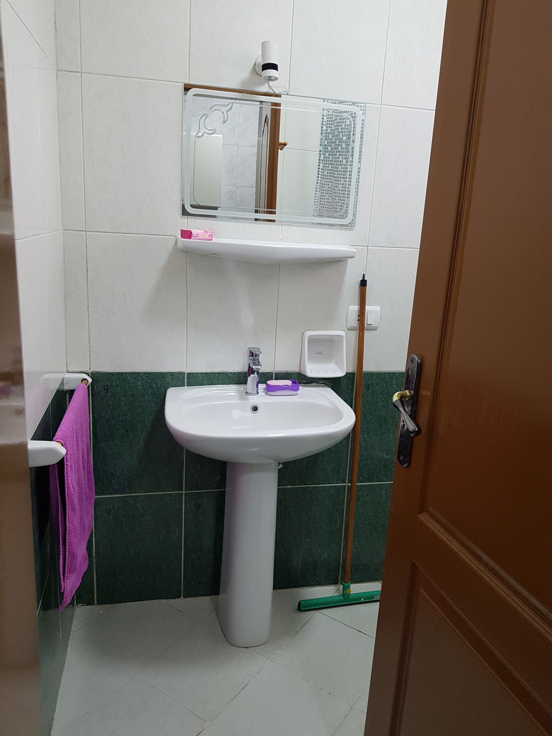 Une salle de bain avec un lavabo blanc, des carreaux verts, un miroir et un balai-serpillière.
