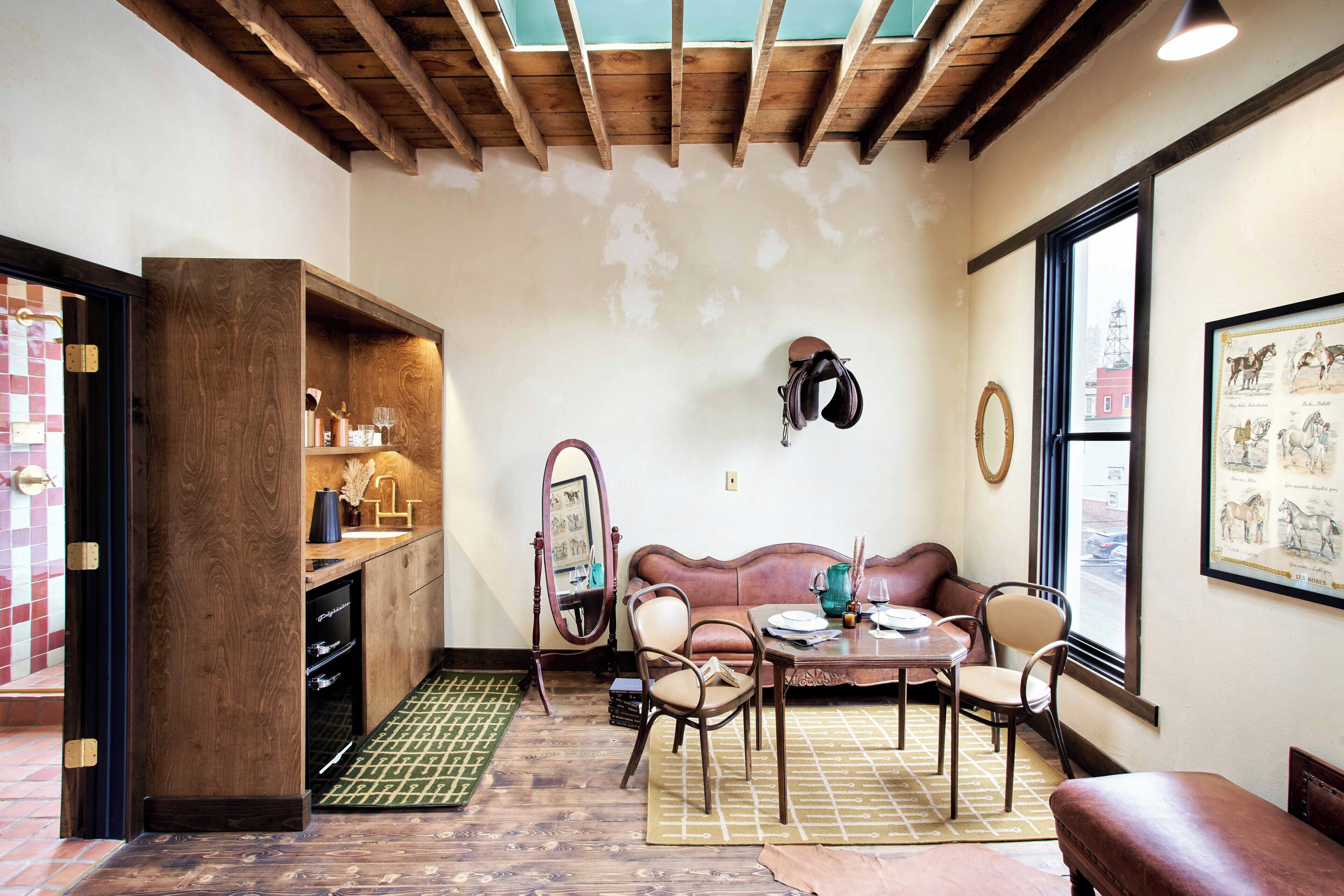 A cozy room with a wooden ceiling, a sofa, a dining table with chairs, a mirror, and a window with a view of a building.