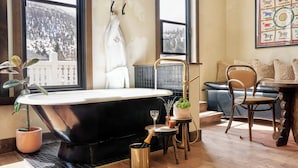 A bathroom with a large bathtub, a potted plant, a champagne bottle, and a view of snow-covered mountains through large windows. - Western Hotel Ouray (Ouray)