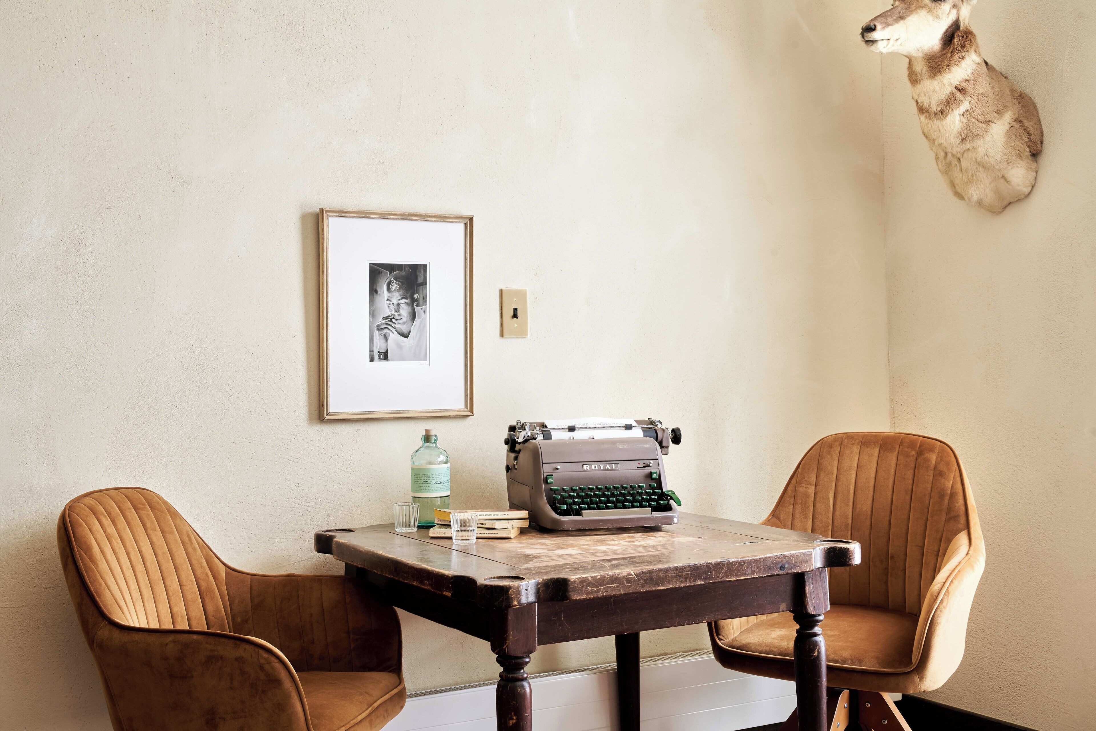 A room with a wooden table, two upholstered chairs, a framed picture on the wall, a typewriter, and a mounted animal head.
