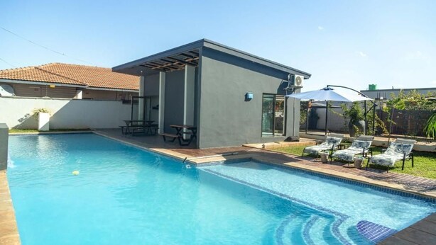 A modern pool area with a covered patio, lounge chairs, and a small table. - Grayville Apartments #5 (Lusaka)