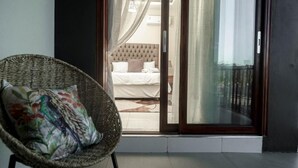 A balcony with a wicker chair and a bed visible through a glass door. - Grayville Apartments #5 (Lusaka)