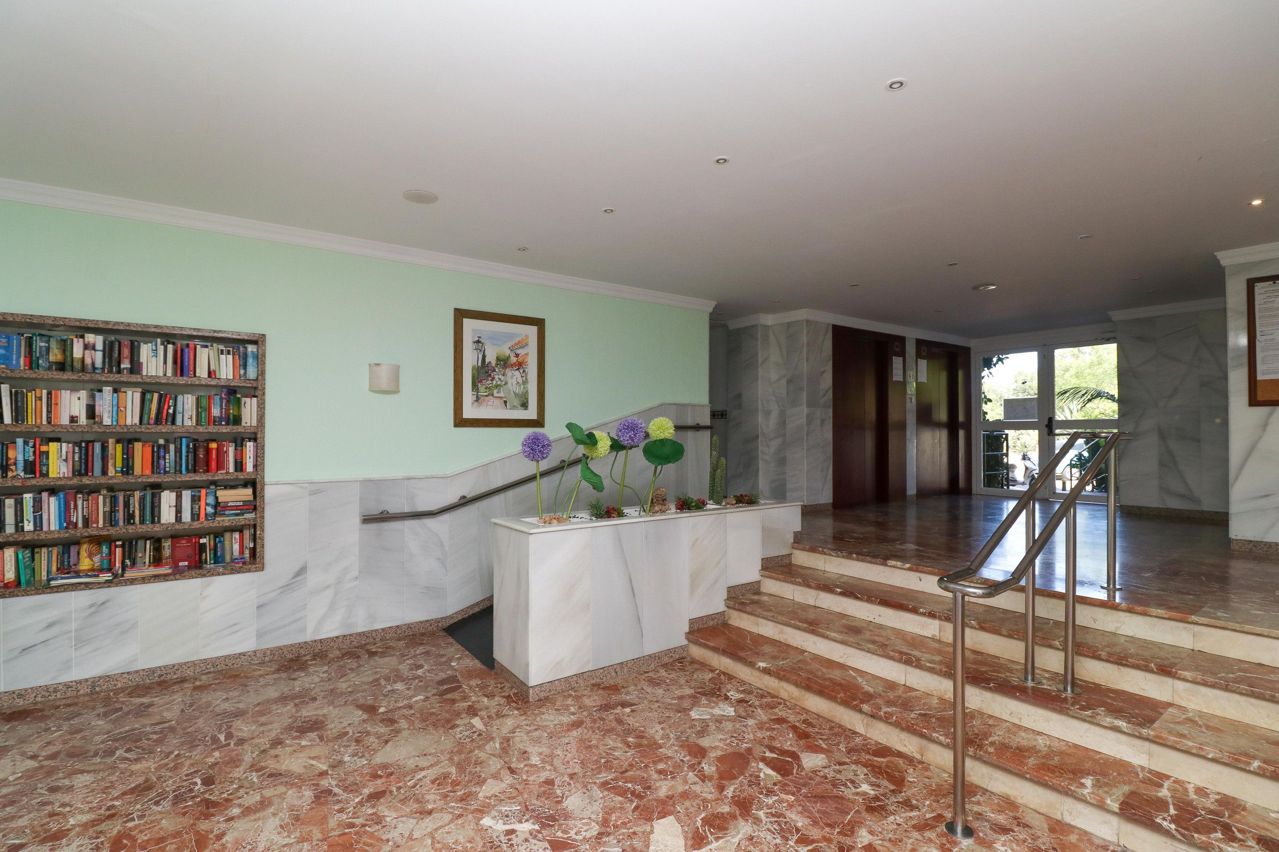 A lobby with a marble staircase, a bookshelf, and a flower arrangement.