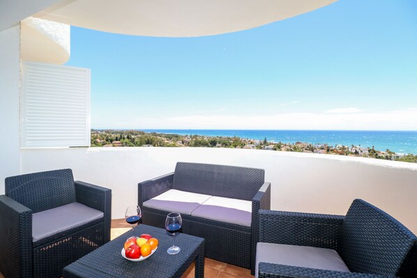 A balcony with wicker furniture, a table with fruits and wine glasses, and a view of the sea and distant landscape. - Renovated studio in Elviria, fantastic sea views (Marbella)