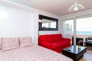 A modern bedroom with a red sofa, a glass coffee table, and a bed with patterned bedding. - Renovated studio in Elviria, fantastic sea views (Marbella)