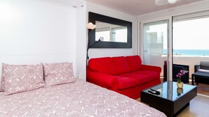 A modern bedroom with a red sofa, a glass coffee table, and a bed with patterned bedding. - Renovated studio in Elviria, fantastic sea views (Marbella)