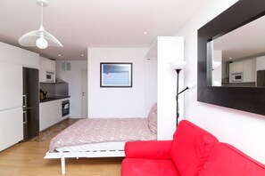 Un dormitorio moderno con cama, un sofá rojo, una lámpara de techo blanca, un televisor montado en la pared y una zona de cocina con electrodomésticos.