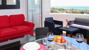 A dining area with a glass table, red and white plates, cutlery, and wine glasses. There is a red sofa and wicker furniture in the background.