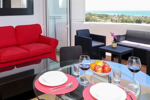 Un comedor con una mesa de vidrio, platos rojos y blancos, cubiertos y copas de vino. Se ve un sofá rojo y muebles de mimbre al fondo.