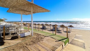 Zona de playa con muelle de madera, sombrillas de paja y sillas reclinables.