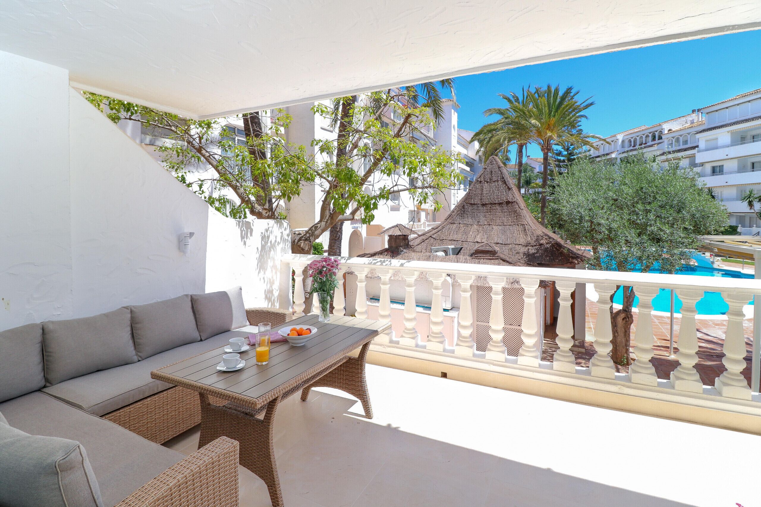 A balcony with a wicker sofa, a table with a bowl of fruit and a glass of orange juice, and a view of a pool and buildings in the background.