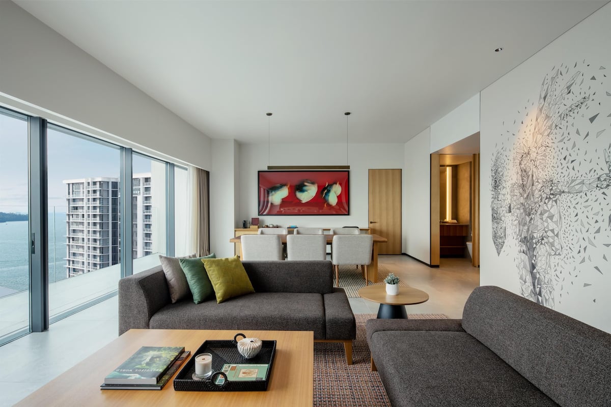 A modern living room with a large window, a sofa, a coffee table, and a dining area with a red abstract painting.