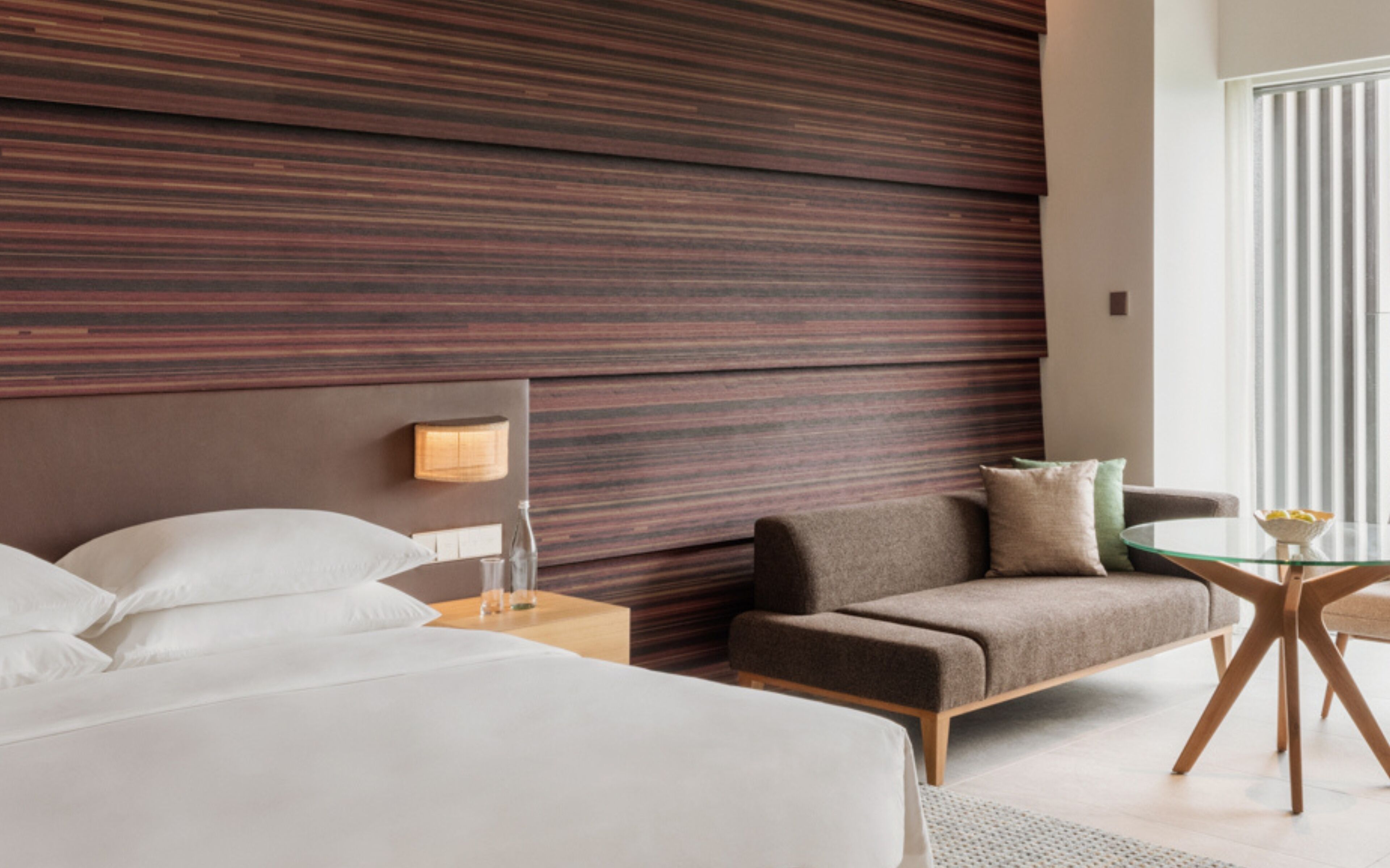 A modern hotel room with a large bed, a sofa, a glass table, and a wall with horizontal wooden panels.