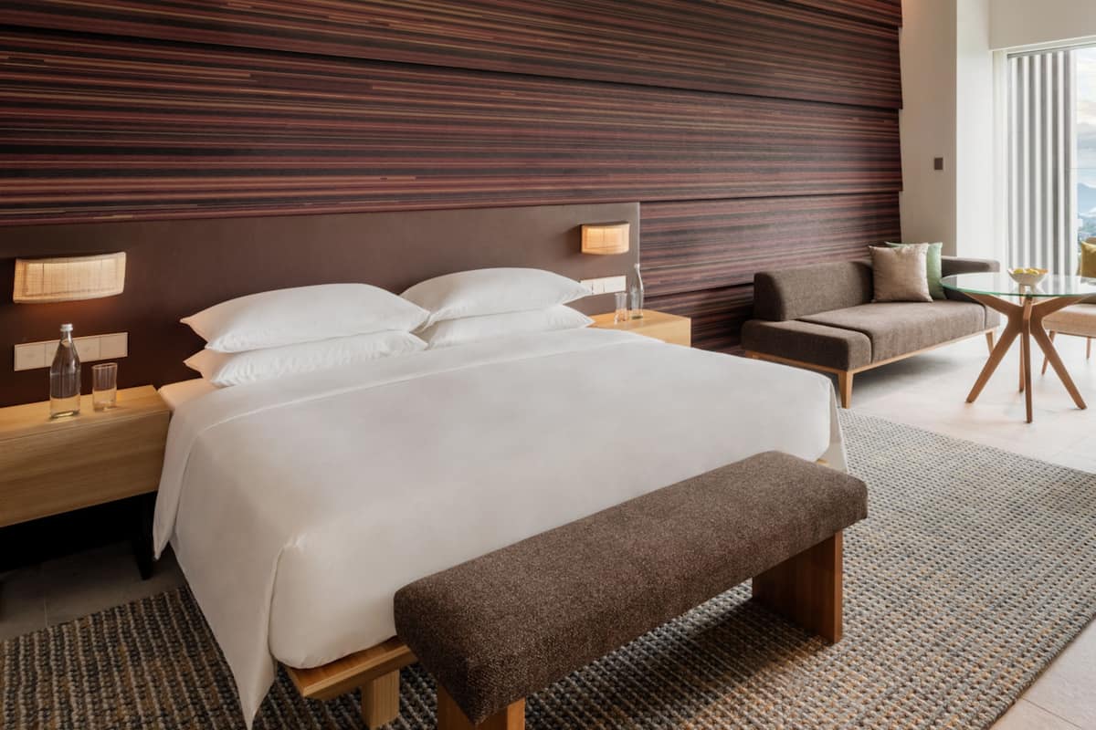A modern hotel room with a large bed, bedside tables, a bench, a chair, and a small table with a lamp.