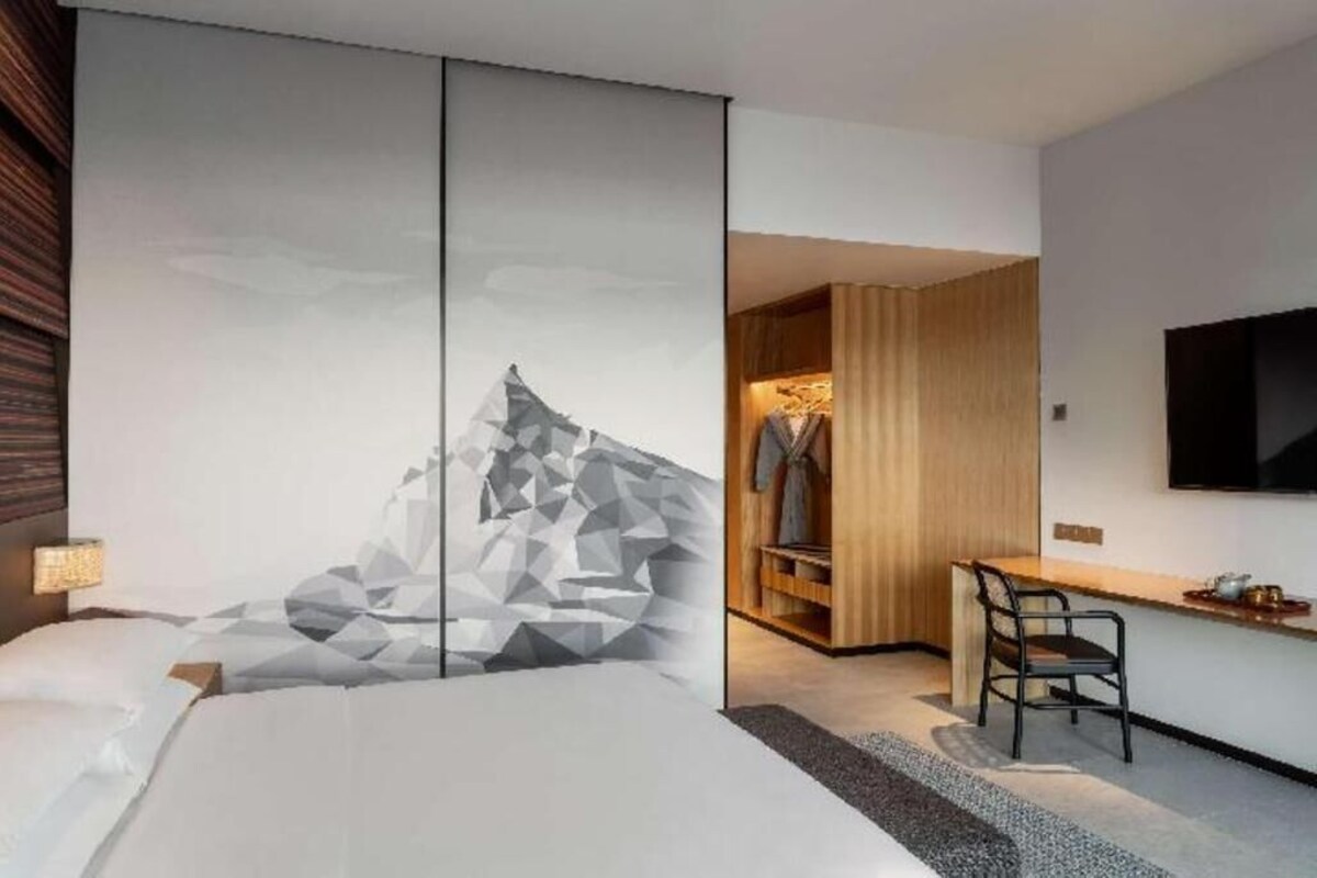 Habitación de hotel moderna con una cama grande, un cuadro de arte geométrico, un armario de madera, un escritorio con silla y un televisor de pantalla plana montado en la pared.