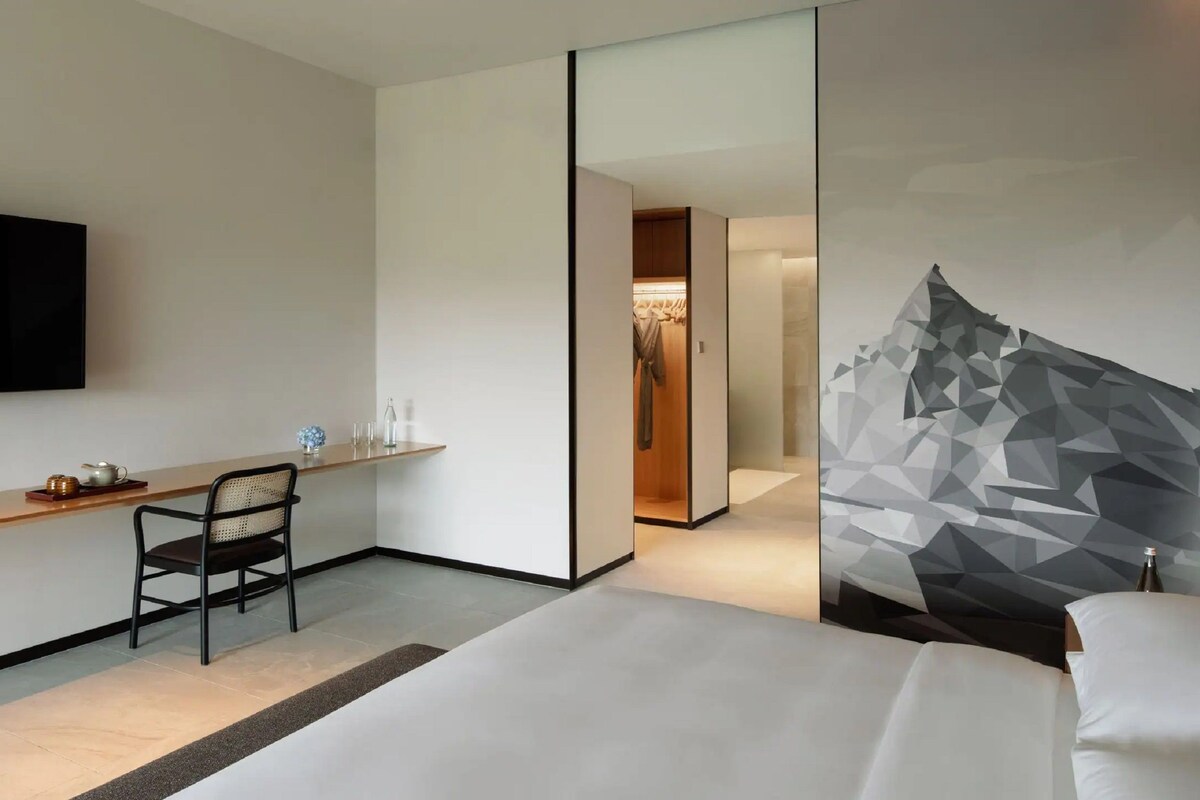 Habitación de hotel moderna con televisión de pantalla plana, escritorio de madera, una silla, una cama y un diseño geométrico en la pared.