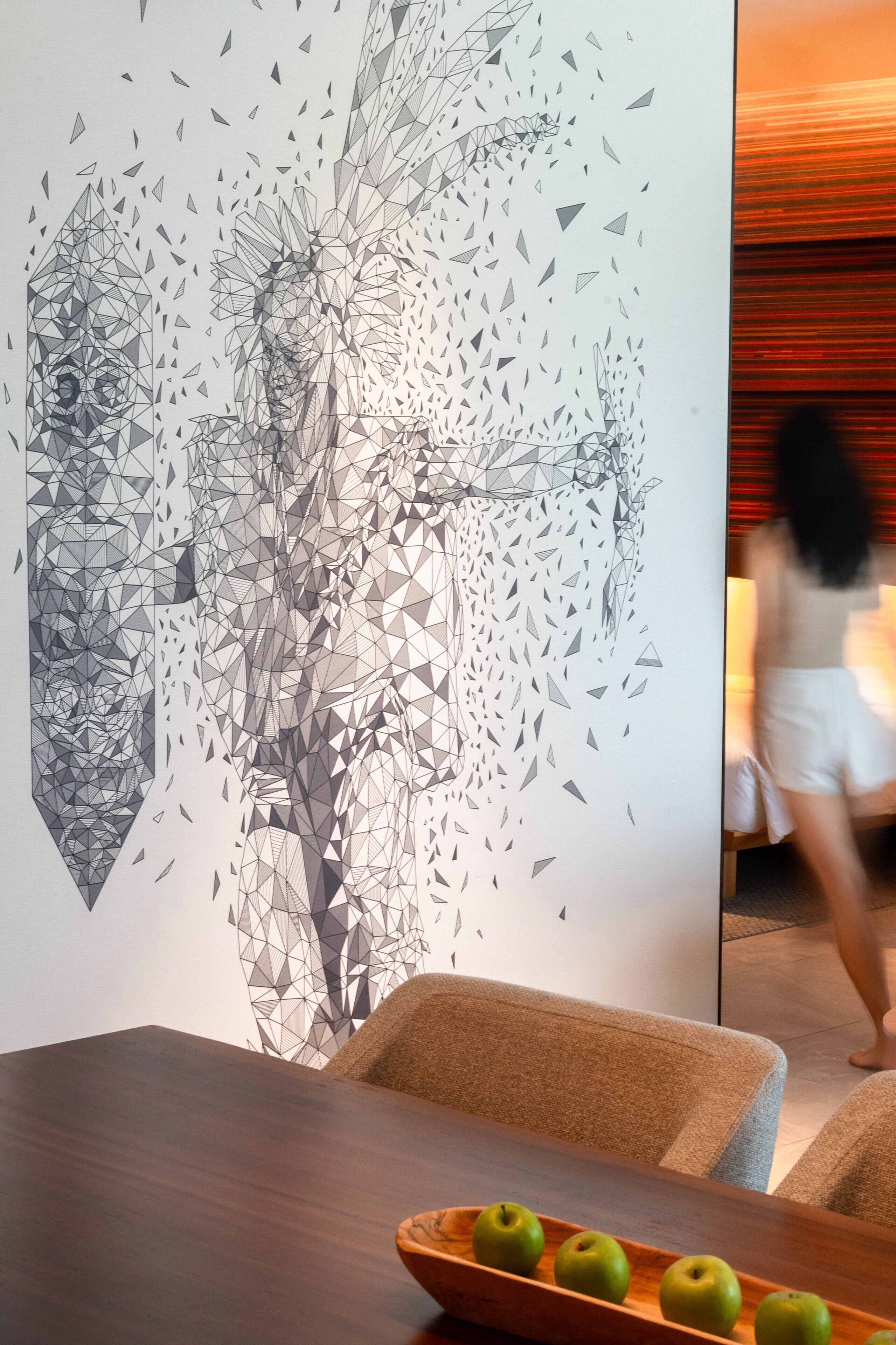 A dining area with a geometric wall art, a wooden table, and a bowl of green apples.