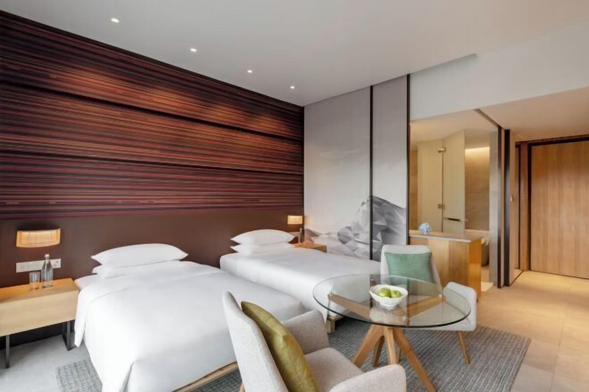 Una habitación de hotel moderna con dos camas, una mesa de vidrio y vista al baño.