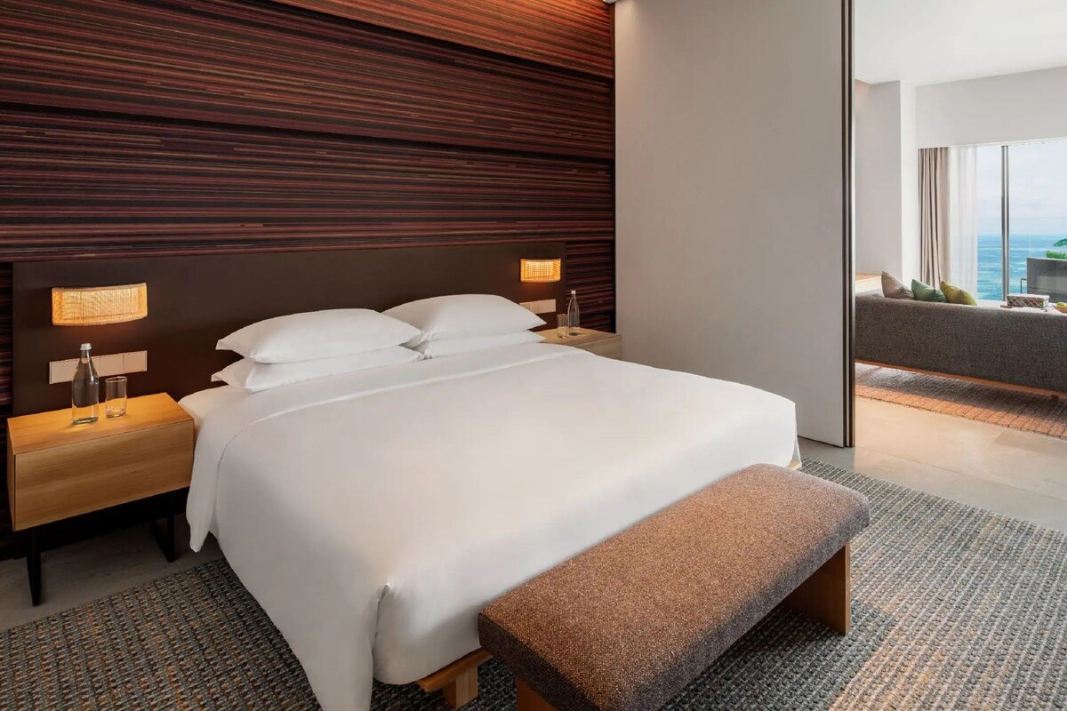 Un hotel moderno con una cama grande, un banco, una mesita de noche y vistas al mar a través de amplias ventanas.