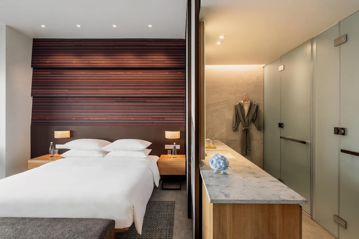 A modern hotel room with a large bed, bedside tables, a wooden headboard, a bathroom with a marble countertop, and a robe hanging on a door.