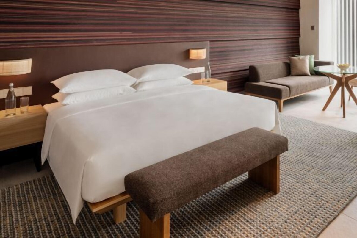 Una habitación de hotel moderna con una cama grande, un banco, un sofá, una mesita pequeña y una lámpara.