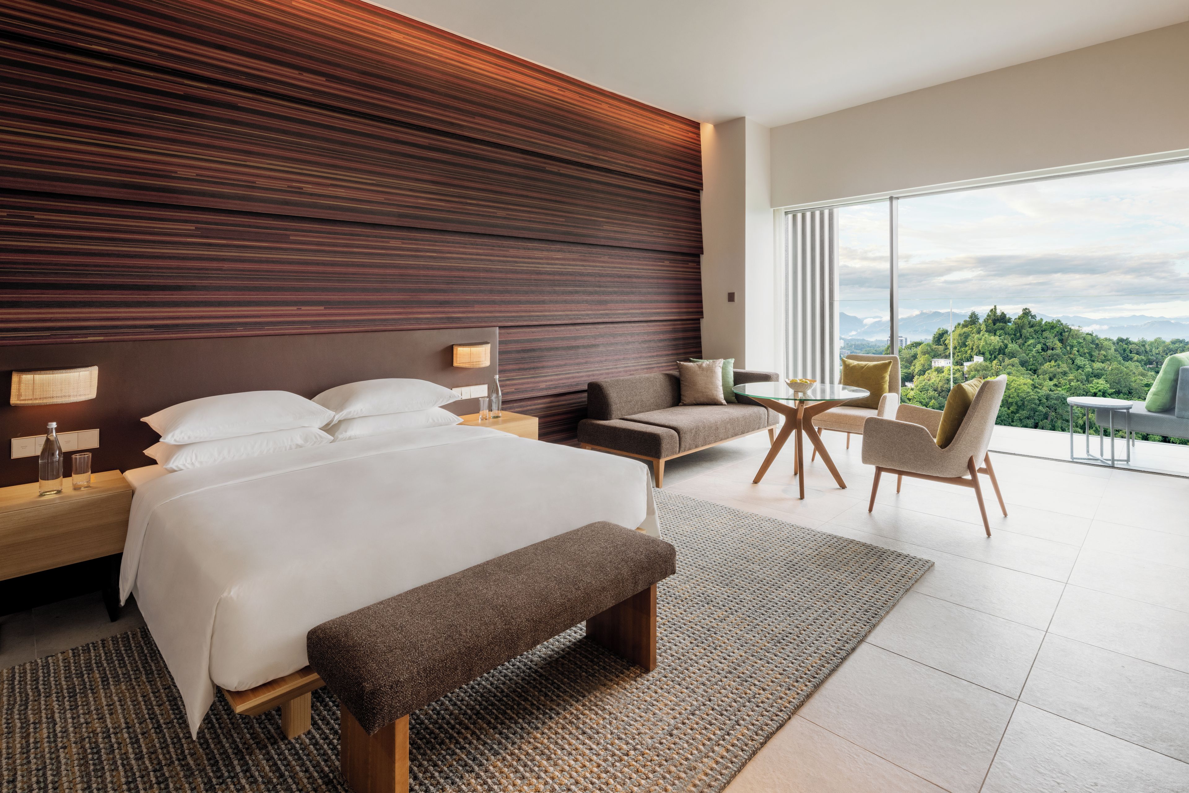 A modern hotel room with a large bed, a sofa, a small table, and a view of the outdoors.