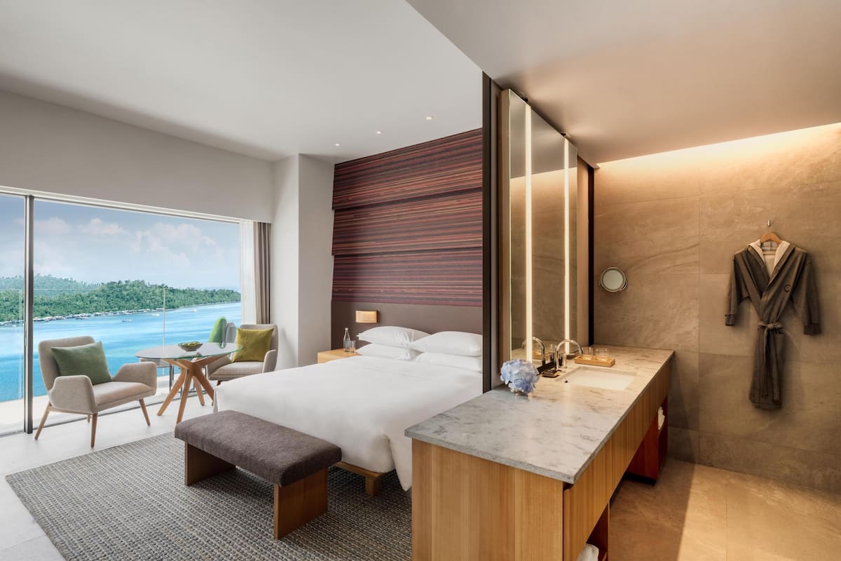 A modern hotel room with a large bed, a desk, a seating area, and a view of the sea.