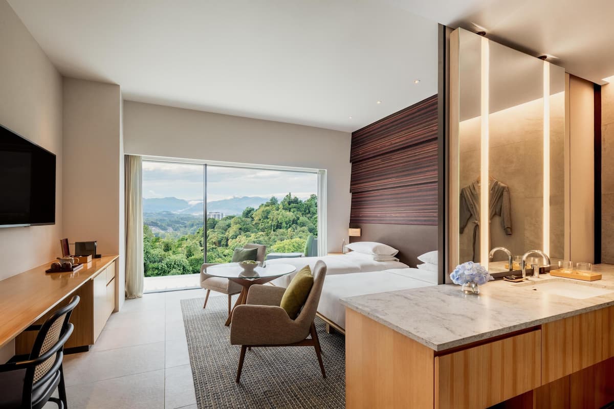 A modern hotel room with a large window, a desk, a chair, a bed, a bathroom with a mirror, and a TV.