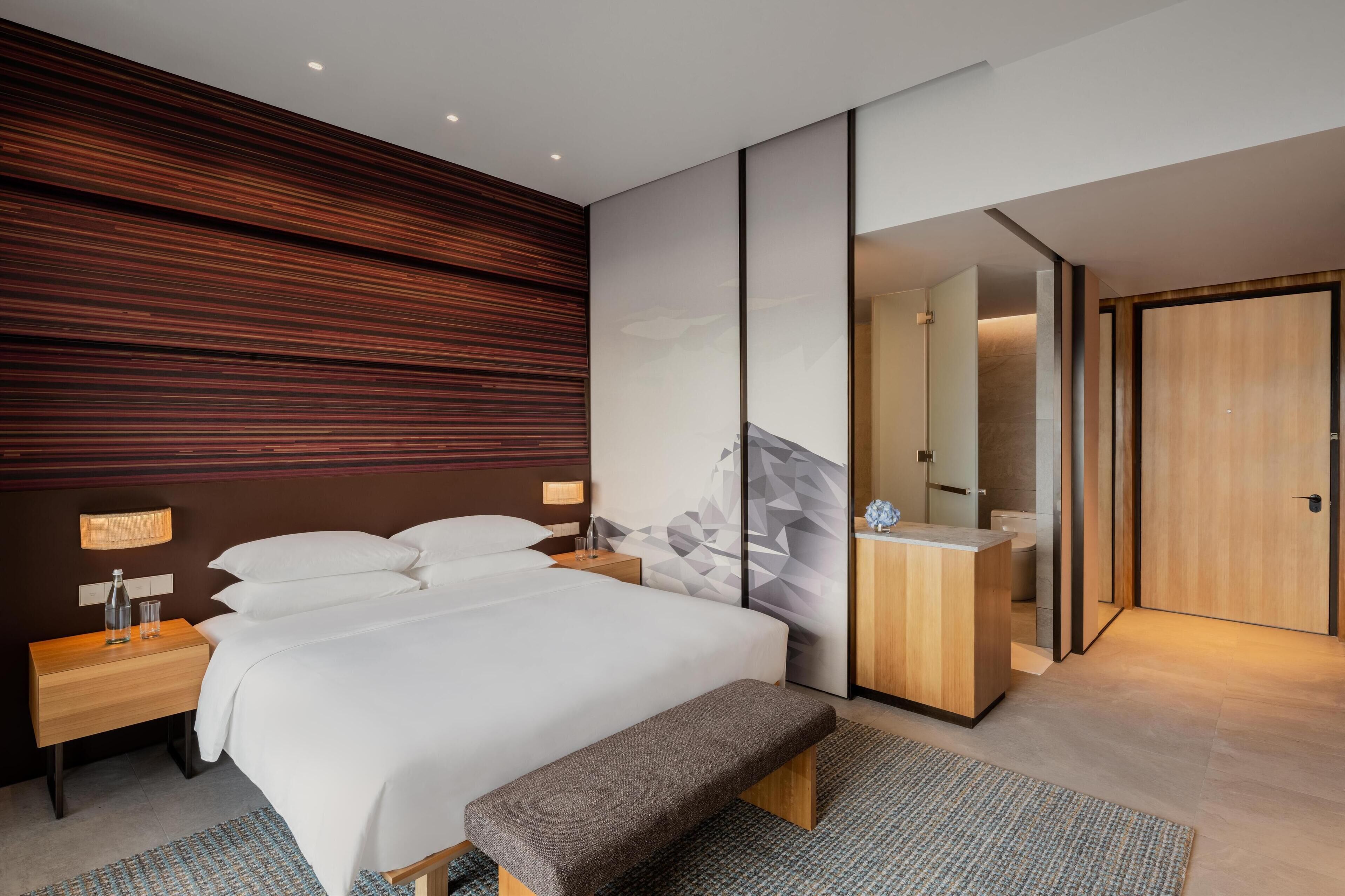 Une chambre d’hôtel moderne avec un grand lit, une banquette, une table de chevet en bois, un miroir et une armoire en bois.