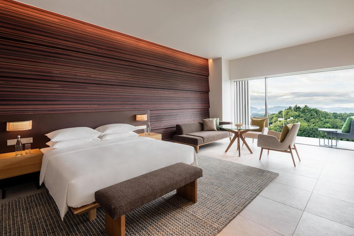 A modern hotel room with a large bed, a sofa, a small table, and a view of the outdoors.