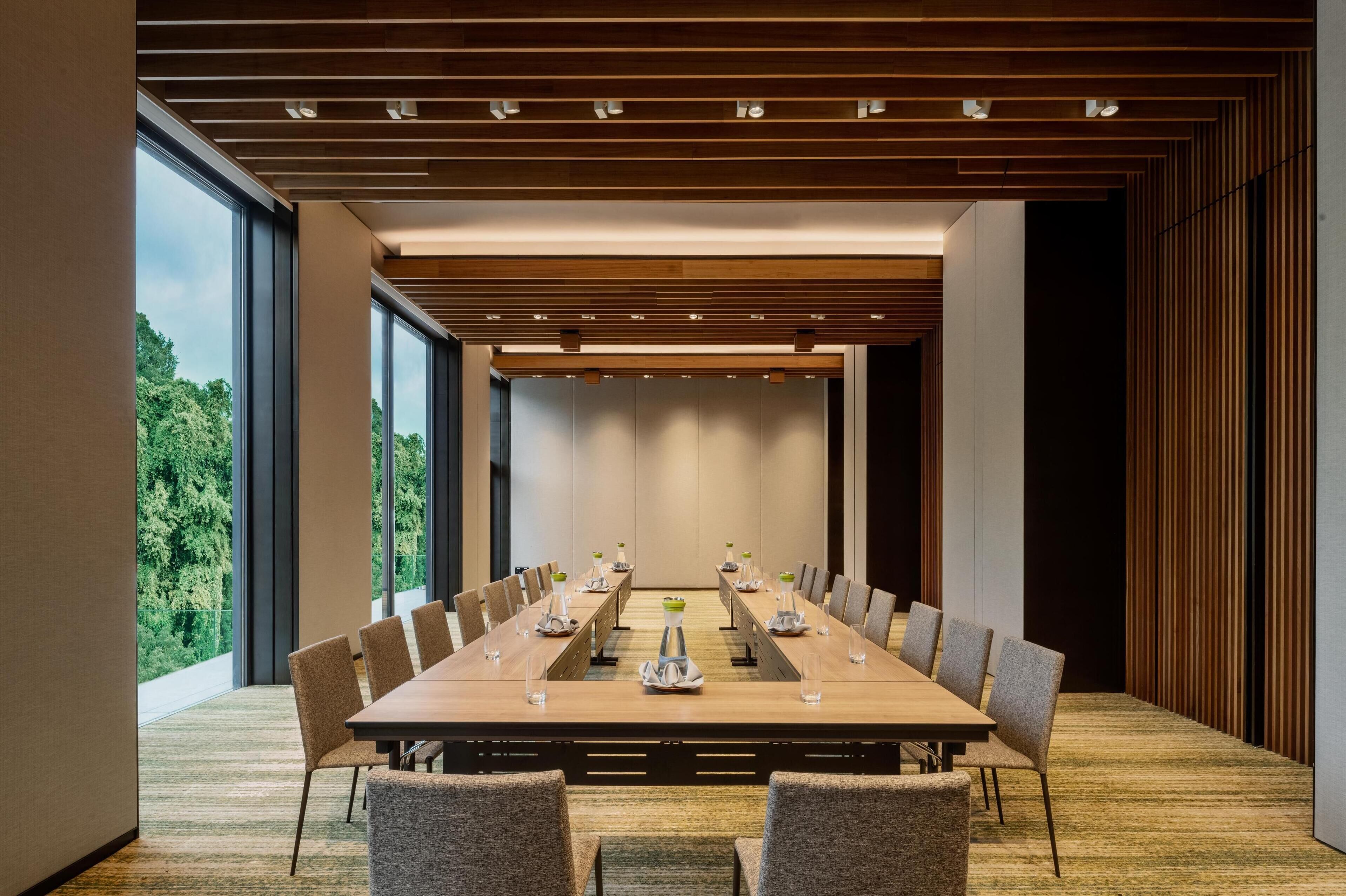 A conference room with a long table, chairs, and a view of greenery outside.