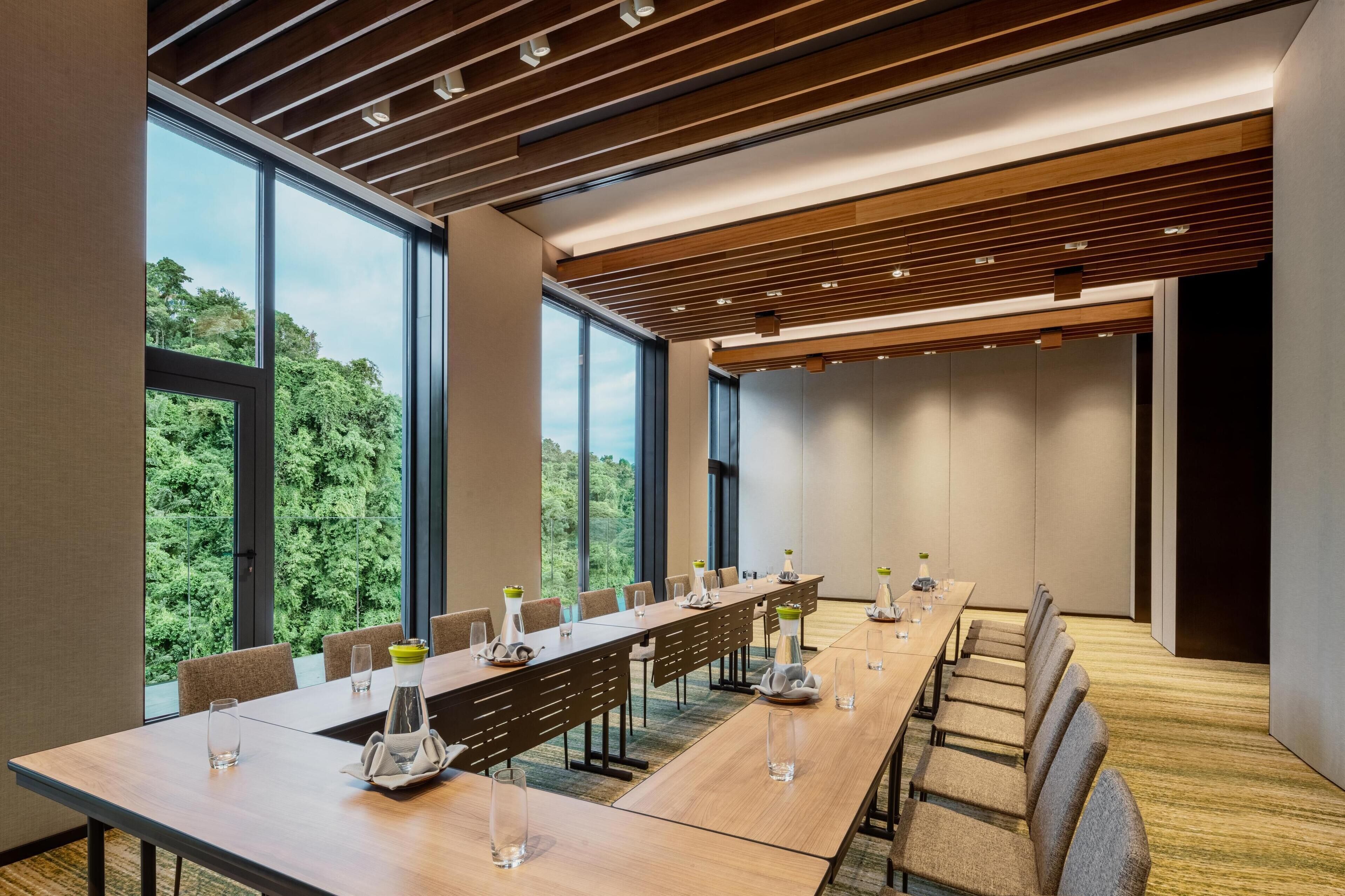 A conference room with a long table, chairs, and large windows offering a view of greenery.