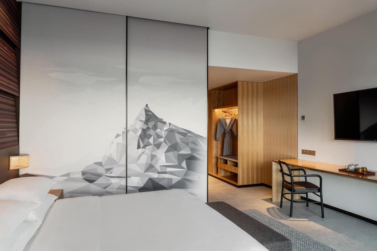 A modern hotel room with a bed, a desk, a chair, a TV, and a wardrobe.