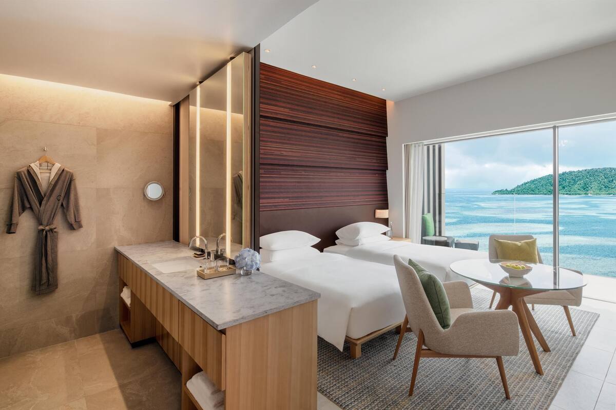 A modern hotel room with a large bed, a desk with a mirror, a small table, and a view of the ocean.