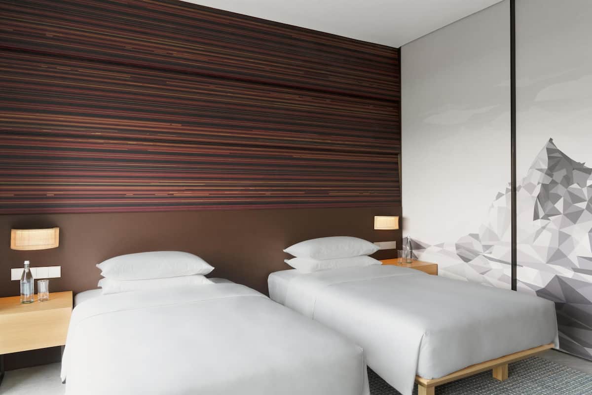 A hotel room with two single beds, a wooden headboard, a nightstand with a lamp, and a geometric wall art.