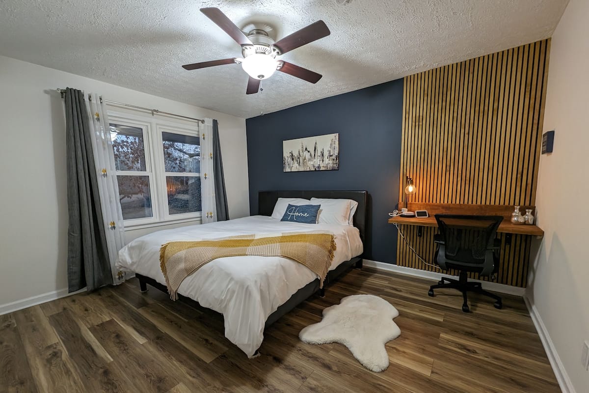 A bedroom with a bed, a desk, a chair, a ceiling fan, a window, and a wall-mounted light.