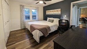 Un dormitorio con una cama, ventilador de techo, ventana con cortinas, un cuadro en la pared y un escritorio con silla.