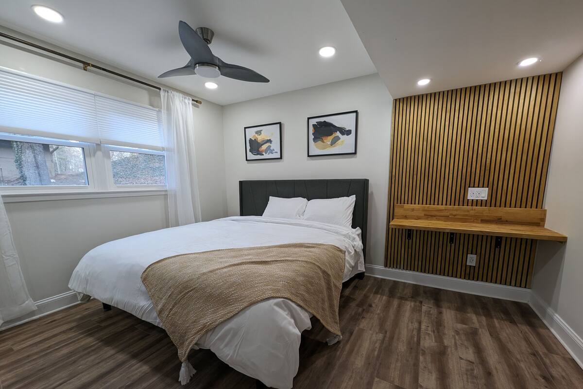 A modern bedroom with a bed, ceiling fan, recessed lighting, wooden panel wall, and artwork.