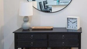 Une console en bois foncé, surmontée d’un miroir rond, agrémentée d ’une lampe blanche, d’une horloge et de deux paniers en osier.