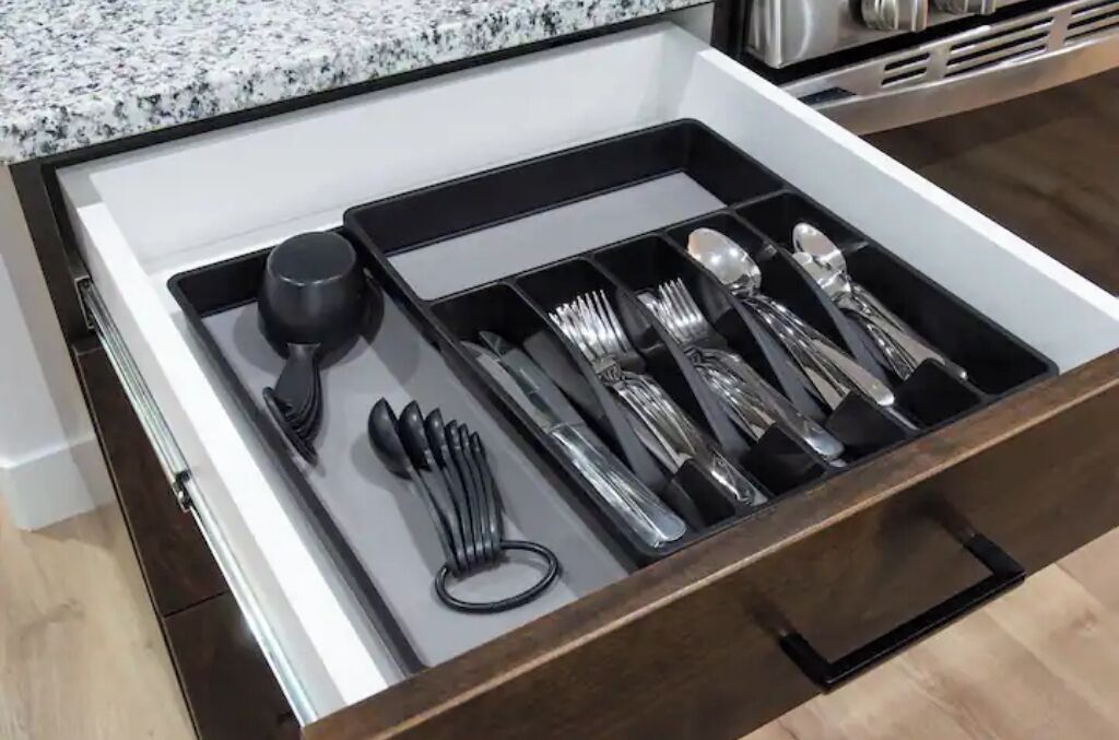 Un tiroir contenant des ustensiles de cuisine rangés avec soin, notamment une louche noire, des fourchettes, des couteaux et des cuillères.