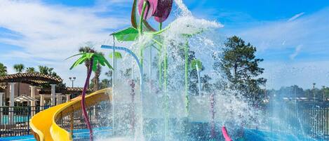 Un parc aquatique avec un toboggan jaune, des jets d’eau et des palmiers.