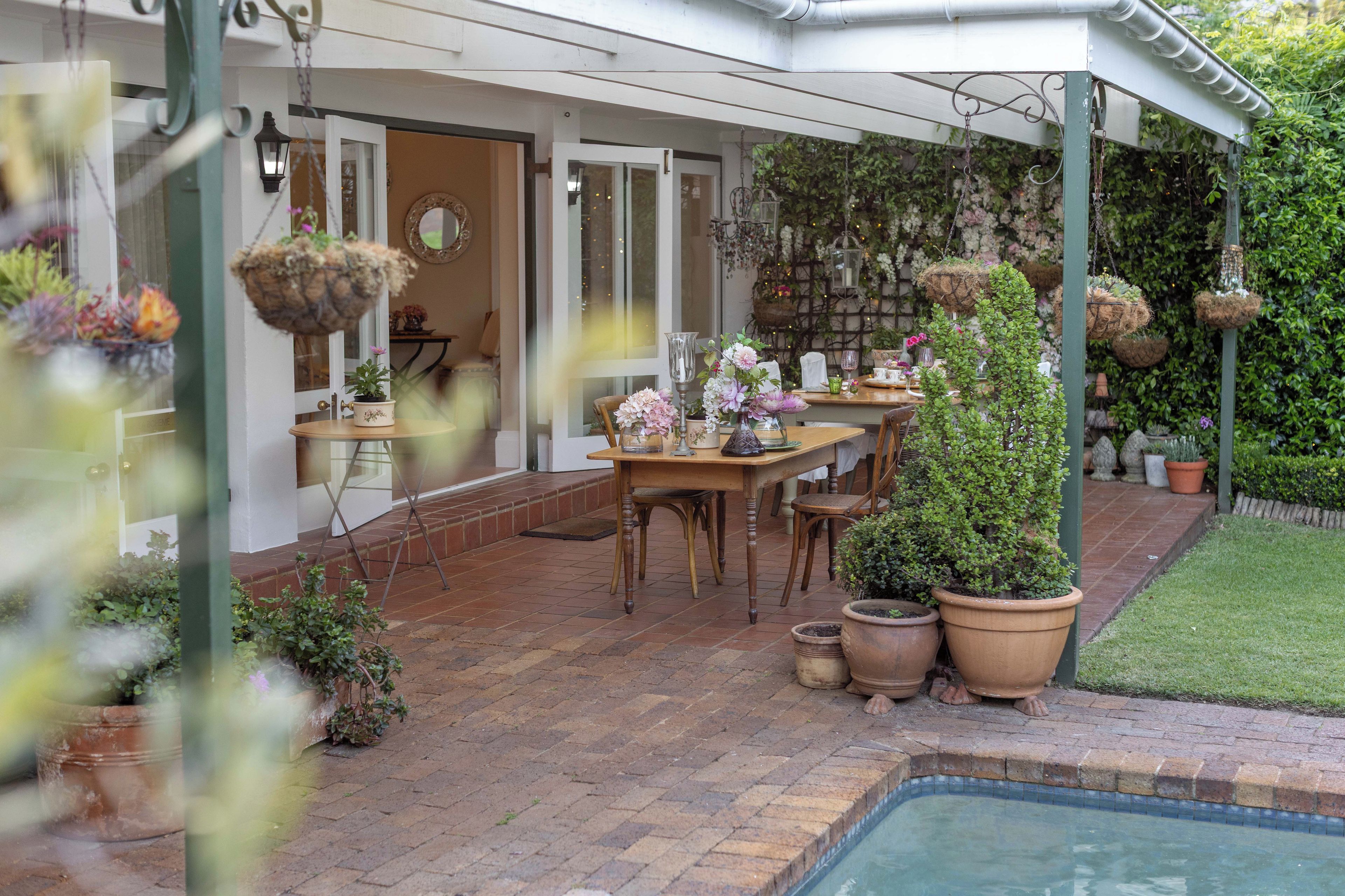 A covered patio area with a pool, a dining table, chairs, hanging planters, and potted plants.