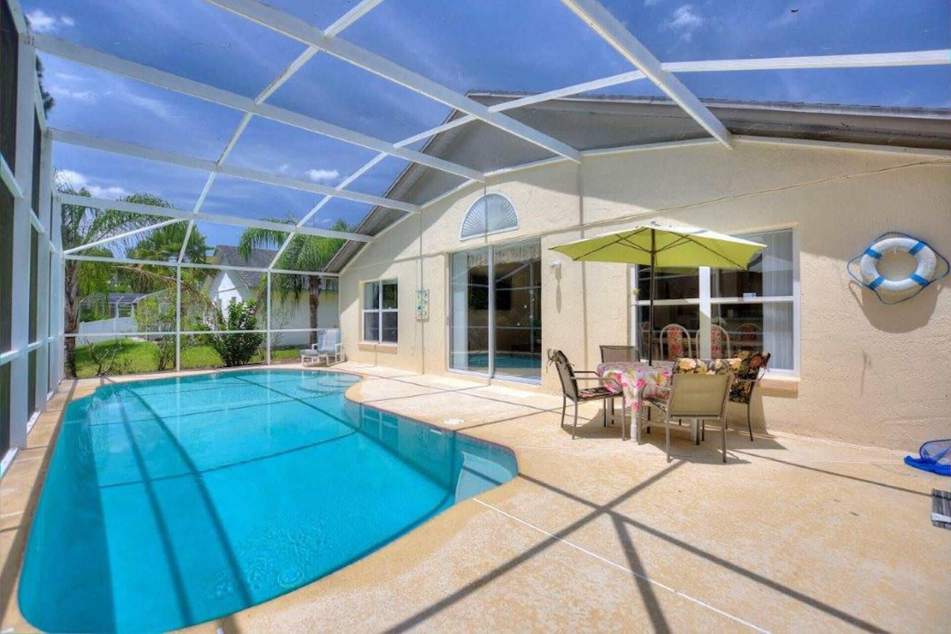 Une piscine extérieure dotée d’une structure en verre, un espace patio avec des chaises et une table, et un anneau de sauvetage fixé au mur.
