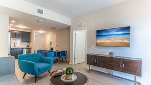 Een moderne woonkamer met een blauwe fauteuil, een eethoek met een tafel en stoelen, een keuken met donkere kastjes en een grote televisie aan de muur.