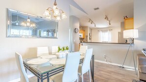 A modern dining area with a glass table and white chairs, a large mirror above, and a kitchen in the background. - 17637 Silver Creek (Clermont)