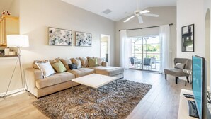 A spacious living room with a beige sofa, a coffee table, and a large rug. There are two ceiling fans, a TV, and artwork on the walls. - 17637 Silver Creek (Clermont)