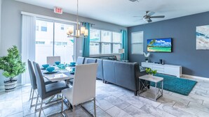 Un salon moderne comprenant un coin repas, des canapés gris, un téléviseur fixé au mur et un ventilateur de plafond.