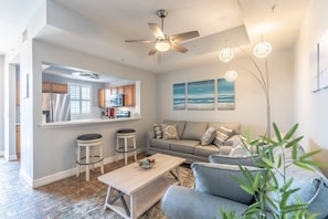 A modern living room with a sofa, coffee table, and bar stools. A kitchen is visible in the background with a refrigerator and stove. - 617 Little Harbor (Ruskin)