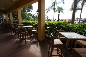 Relax in a tranquil outdoor setting with tables and chairs under a covered roof, surrounded by lush greenery and palm trees.